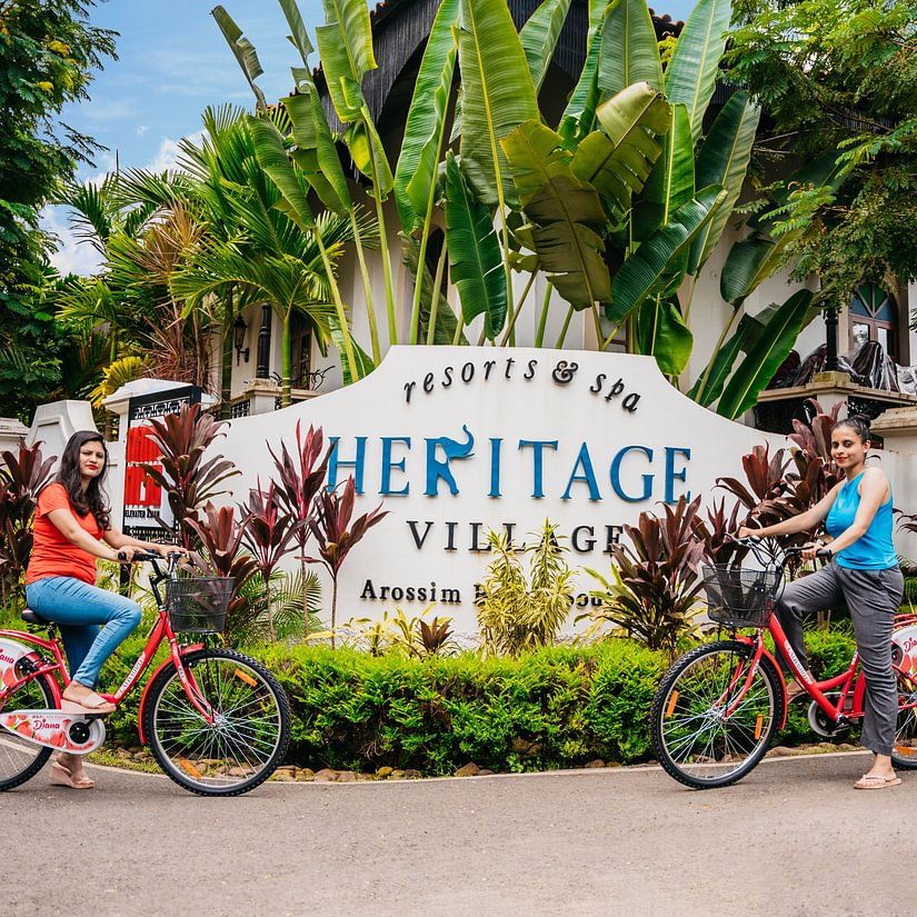 two women on bicycles posing against the backdrop of Heritage Village Resorts & Spa, Goa's sigange