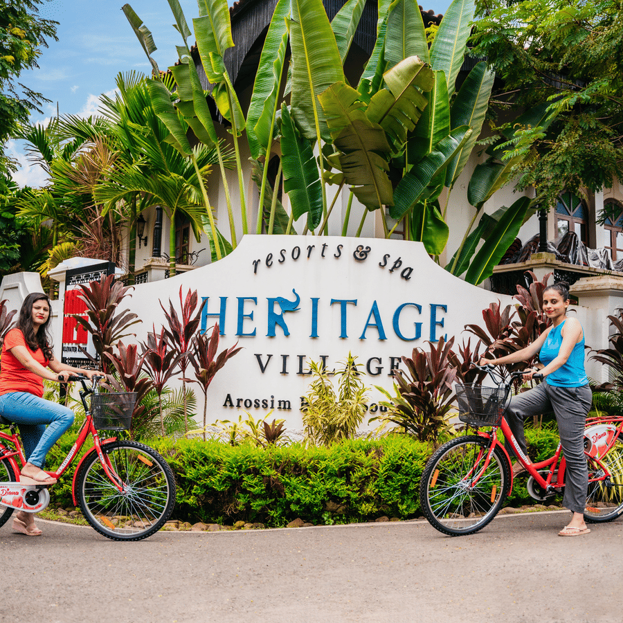 Guests riding bicycles past the main entrance signage of Heritage Village Resorts & Spa, Goa.