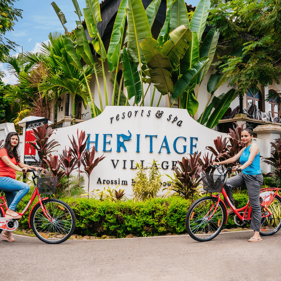 Guests riding bicycles past the main entrance signage of Heritage Village Resorts & Spa, Goa.