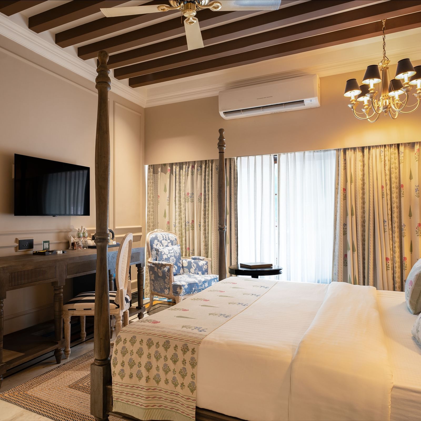Elegant hotel room with a four-poster bed, floral curtains, a chandelier, and wooden ceiling beams. The room features a wall-mounted TV, a wooden desk with chairs, and a blue floral armchair by large windows with sheer drapes - Panna Vilas Palace