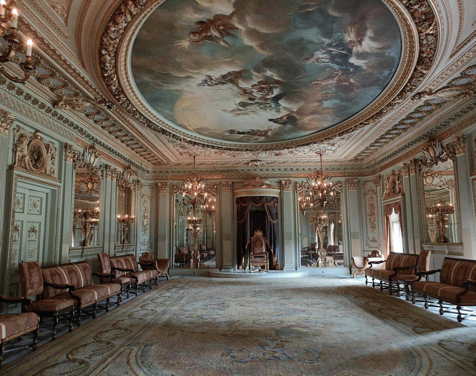 Highly ornate room with a painted ceiling of a mythological scene, gold trim, chandeliers, and rows of velvet seating leading to a central throne under a canopy.
