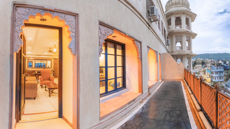 Curved exterior corridor of the best hotel in Nathdwara, Shri Vallabh Vilas Lords Plaza, with intricately carved windows and a view of the nearby city and lake in the backdrop.