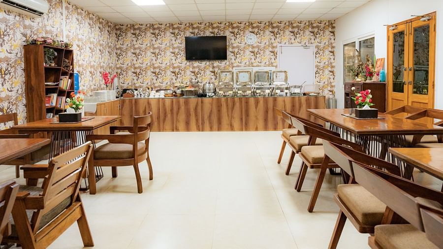 Aisle in the dining area at Neer Ganga, featuring wooden-furnished tables and chairs, a buffet area, and a shelf stocked with kitchen items, creating a cozy and well-equipped dining space.