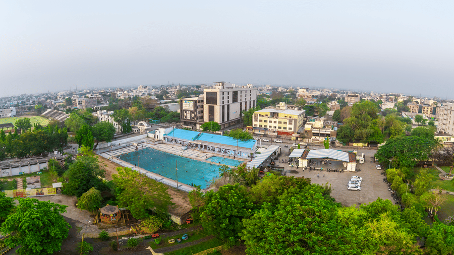  An aerial view of the Nanded city featuring a large outdoor swimming pool, lush green trees, and the hotel building under a clear sky - Skkylight Lords Inn, Nanded