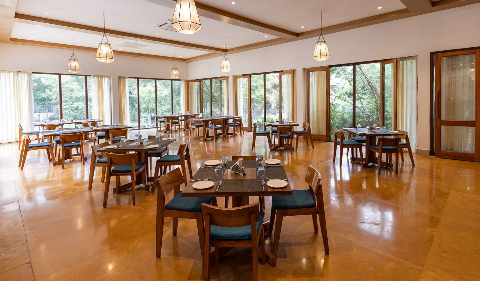Seating arrangement in Azrak Restaurant at Saraca Resort & Spa Corbett with wooden flooring and wooden panelling on the ceiling alongside large French windows bringing in lots of light