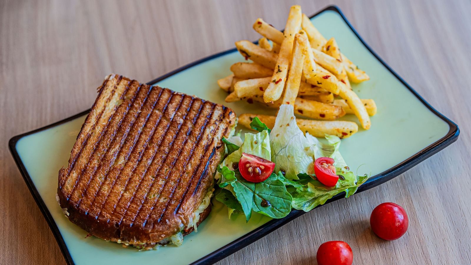 A rectangular plate with a sandwich and vegetables on the side, accompanied by French fries and mini tomatoes placed beside the plate at Indie Stays Mumbai International Airport.