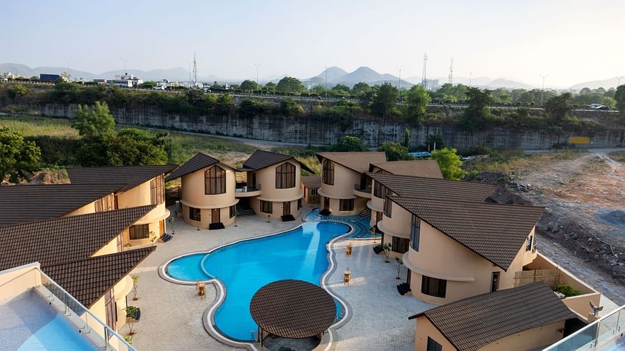 An aerial view of The Qualia Resort Club & Brewery, Udaipur during the daytime, featuring the resort's architecture, swimming pool, and surrounding landscape.