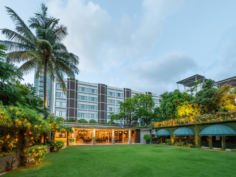Facade view of Kenilworth Hotel with the banquet hall in Park Street, Kolkata, featuring a lawn area, potted plants having fairy lights on it, and a trees on either side of the building.