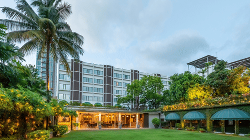 Facade view of Kenilworth Hotel with the banquet hall in Park Street, Kolkata, featuring a lawn area, potted plants having fairy lights on it, and a trees on either side of the building.