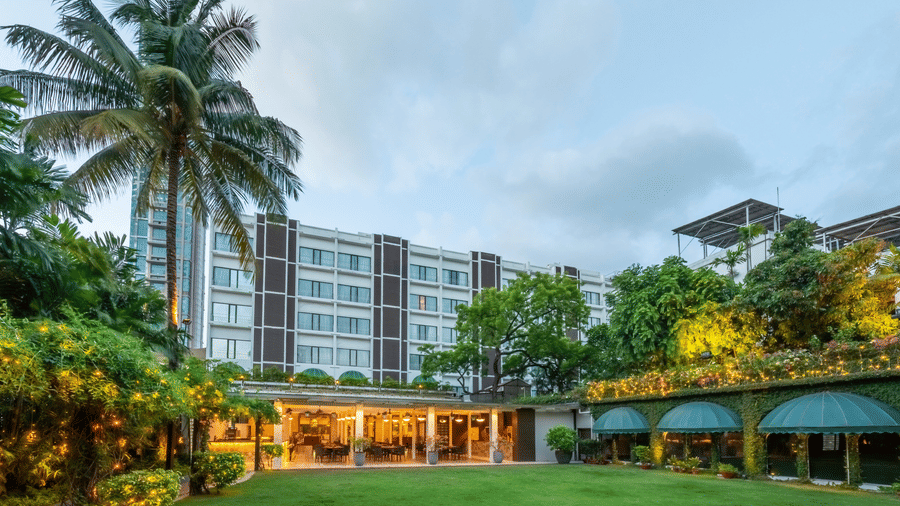 Facade view of Kenilworth Hotel with the banquet hall in Park Street, Kolkata, featuring a lawn area, potted plants having fairy lights on it, and a trees on either side of the building.