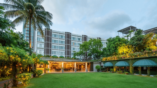 Facade view of Kenilworth Hotel with the banquet hall in Park Street, Kolkata, featuring a lawn area, potted plants having fairy lights on it, and a trees on either side of the building.