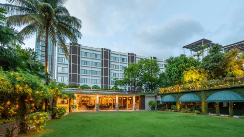 Facade view of Kenilworth Hotel with the banquet hall in Park Street, Kolkata, featuring a lawn area, potted plants having fairy lights on it, and a trees on either side of the building.