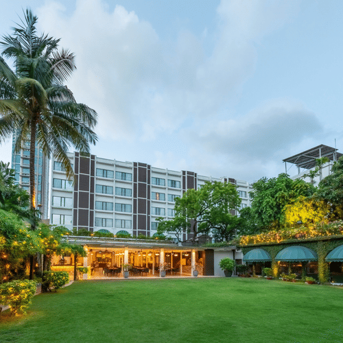 Facade view of Kenilworth Hotel with the banquet hall in Park Street, Kolkata, featuring a lawn area, potted plants having fairy lights on it, and a trees on either side of the building.