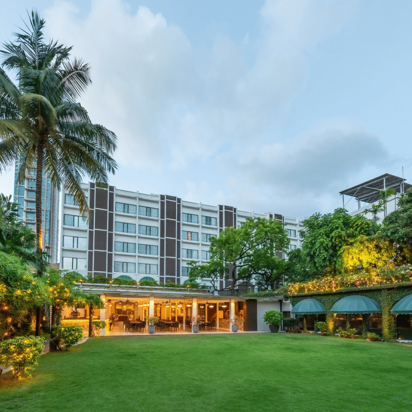 Facade view of Kenilworth Hotel with the banquet hall in Park Street, Kolkata, featuring a lawn area, potted plants having fairy lights on it, and a trees on either side of the building.