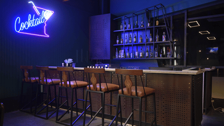 A bar counter with a glowing neon cocktail sign and high stools at Hotel Sonar Bangla, Mayapur.