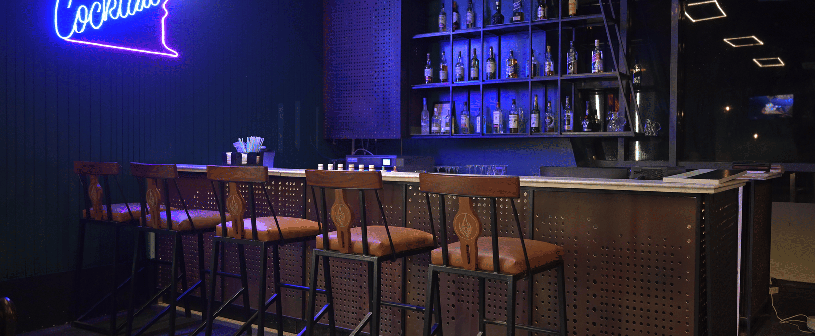 A stylish bar counter with a glowing neon cocktail sign and high stools in an evening setting at Hotel Sonar Bangla Mayapur.