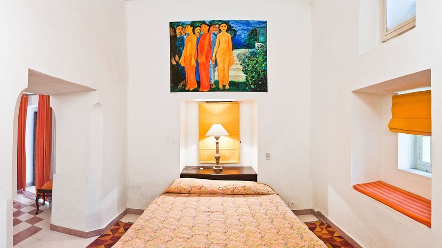 A bedroom with a bed, a lamp, a painting, and a window in Tijara Fort-Palace - 19th Century, Alwar.