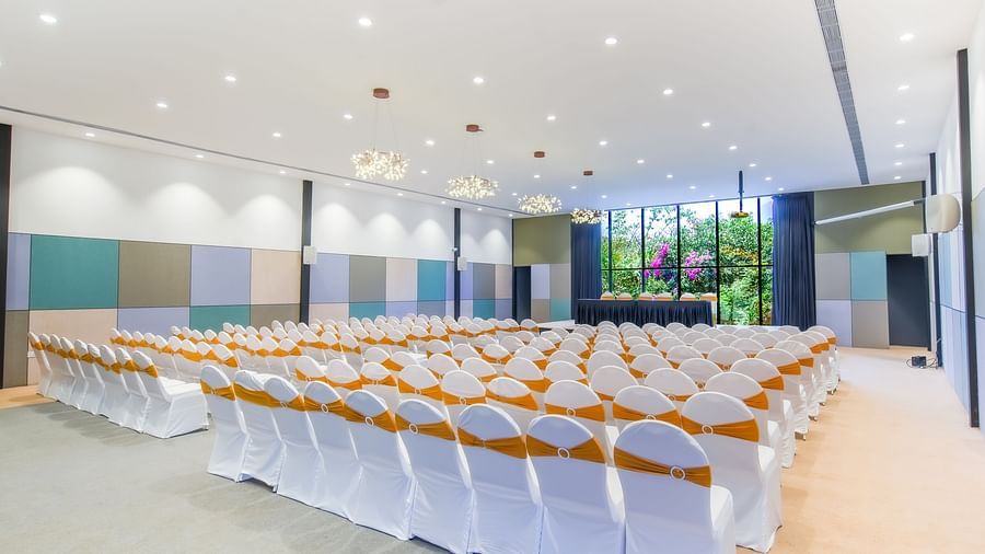 Royal Affair at Darza Resort is arranged with numerous white chairs adorned with orange sashes, facing a stage with the view of flowers in the trees outside.