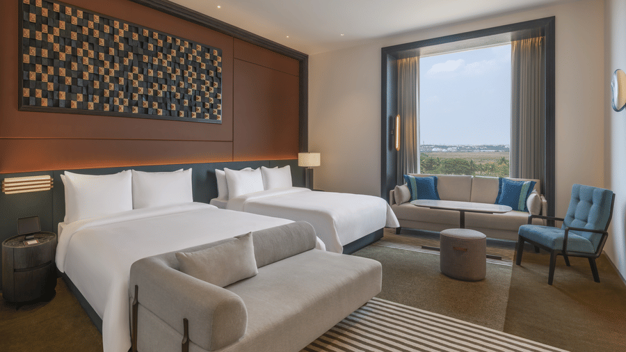 Premium Family Room at The Residency Signature Madurai featuring two double beds, a small gray sofa, a window seating area, and a large decorative wall panel.