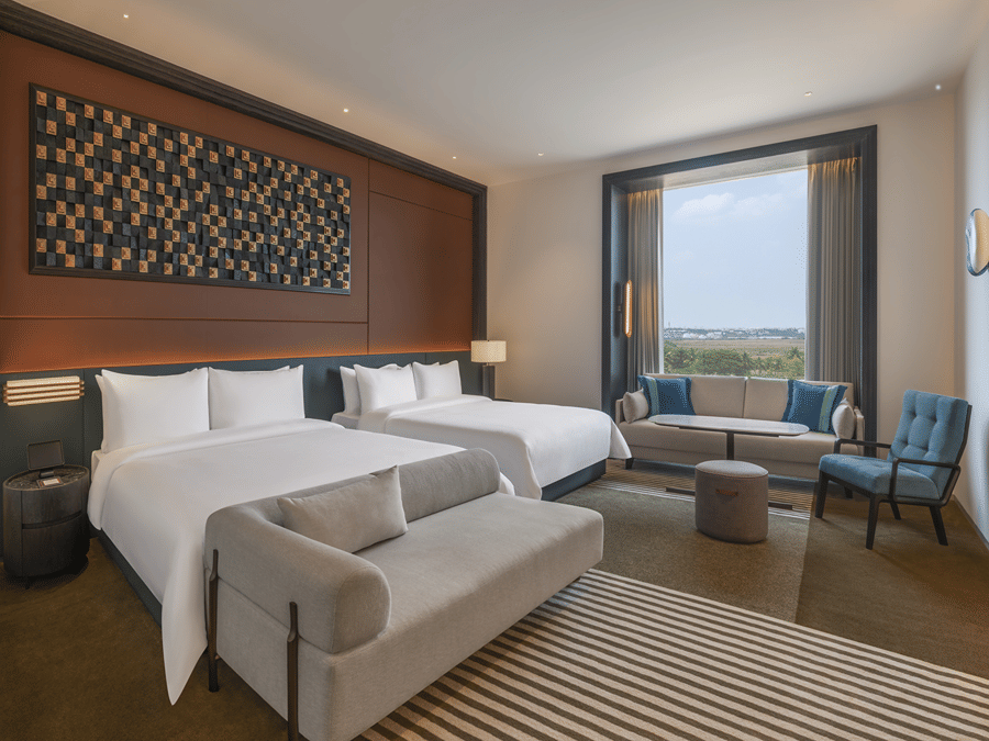 Premium Family Room at The Residency Signature Madurai featuring two double beds, a small gray sofa, a window seating area, and a large decorative wall panel.