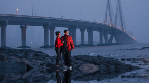 Two people in bright coloured jackets standing on a rocky bank of a river with a bridge in the background.