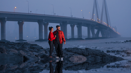 Two people in bright coloured jackets standing on a rocky bank of a river with a bridge in the background.