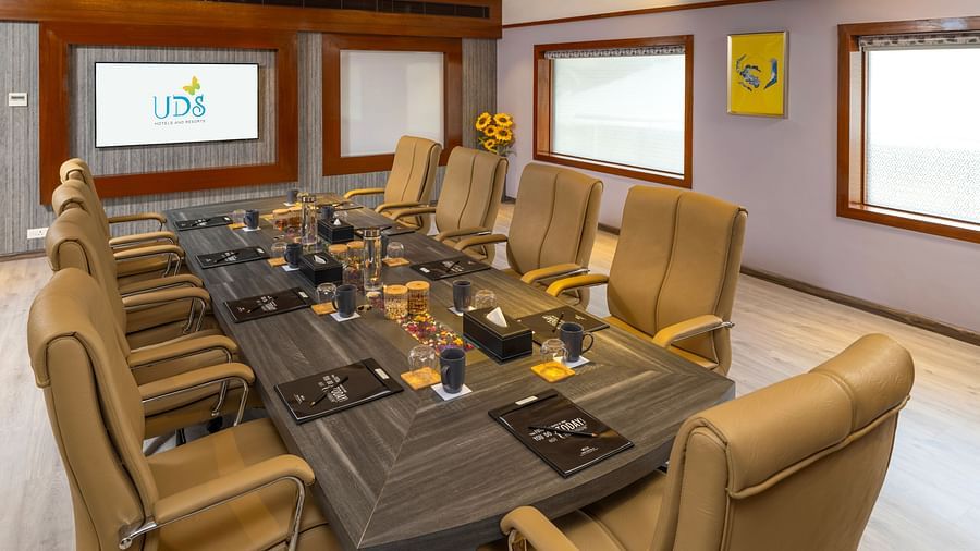 A side view of a boardroom featuring chairs around a table with a mug, notepad, pen, and water glass on the surface, along with a presentation screen at the front - Uday Samudra Leisure Beach Hotel