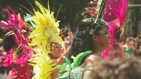 Carnival participants wearing vibrant feathered headdresses and elaborate costumes celebrate outdoors, surrounded by crowds in festive atmosphere