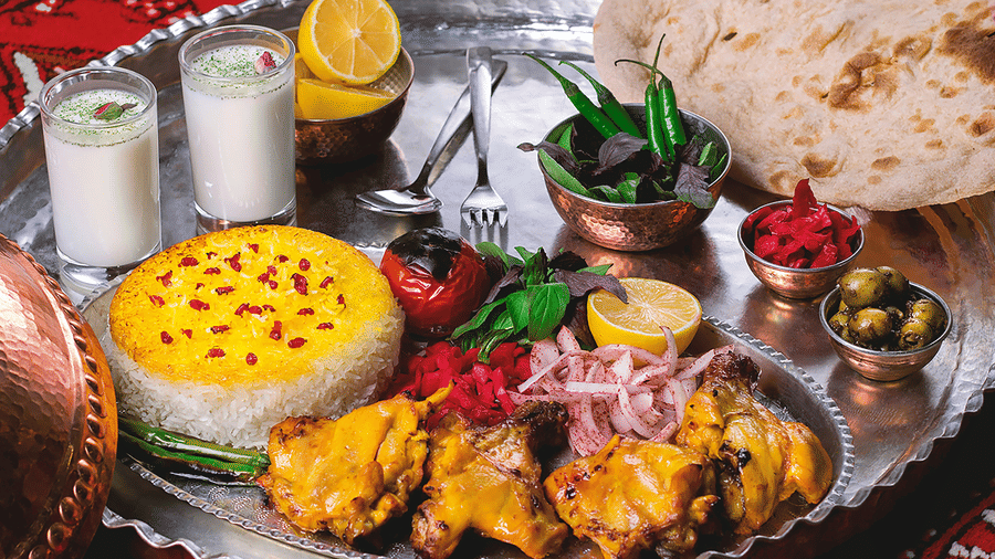 An elaborate and colourful Thali featuring yellow rice, dahi vada (lentil dumplings in yoghurt), grilled chicken pieces, and various fresh garnishes.
