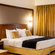 Hotel room at Featherlite Evoma Hotel with a bed dressed in white and gold linen, a wooden headboard and long golden curtains close by.