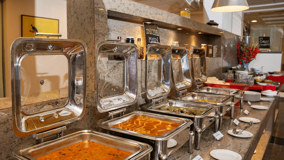 Stainless steel chafing dishes with open lids serving hot curries and gravies buffet-style at The Courtyard Restaurant, Featherlite Evoma Hotel.