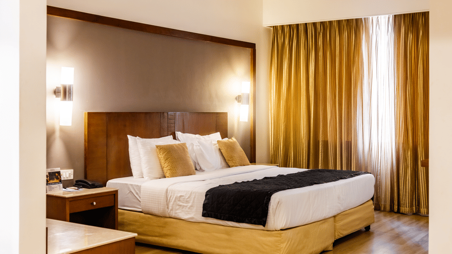 Hotel room at Featherlite Evoma Hotel with a bed dressed in white and gold linen, a wooden headboard and long golden curtains close by.