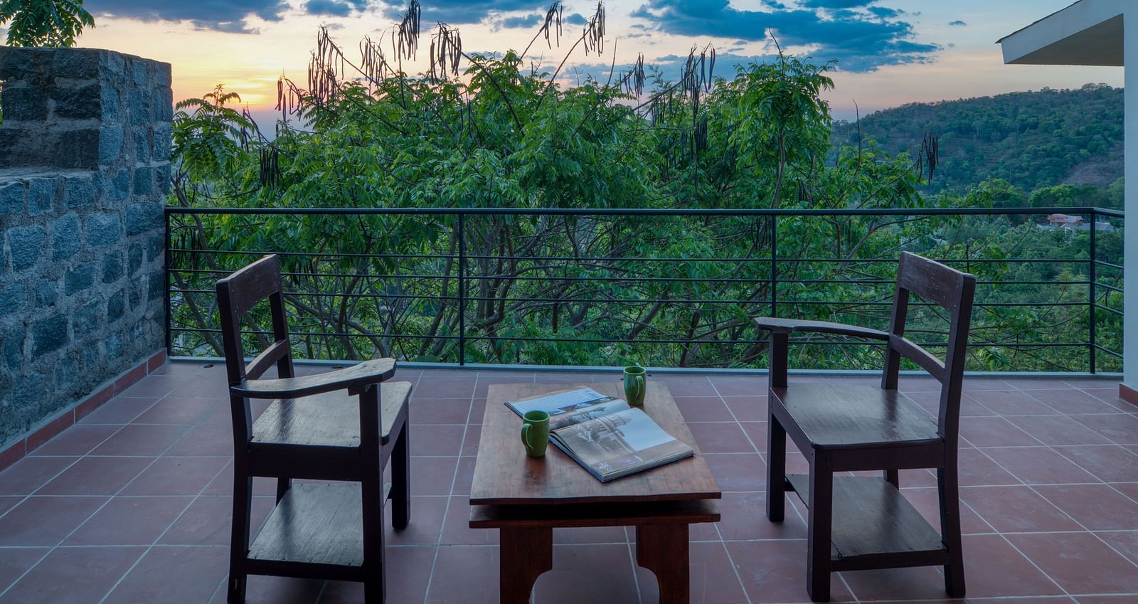 A serene balcony with two wooden chairs and a small table, overlooking a lush green landscape and a beautiful sunset.  | Mango Hill, Yercaud