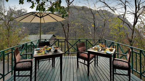 Outdoor wooden deck with 2 set dining tables and an umbrella, overlooking a forested mountain landscape | Neemrana's Glasshouse On The Ganges - 21st Century, Rishikesh