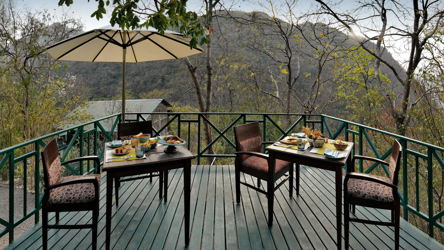 Wide wooden terrace with dining furniture and a large sun umbrella overlooking the dense forest and valley views at Neemrana’s Glasshouse on The Ganges offering a beautiful location for guests to enjoy their breakfast or tea while surrounded by nature