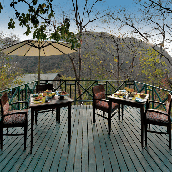 Wide wooden terrace with dining furniture and a large sun umbrella overlooking the dense forest and valley views at Neemrana’s Glasshouse on The Ganges offering a beautiful location for guests to enjoy their breakfast or tea while surrounded by nature