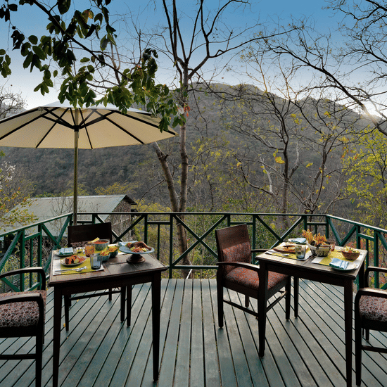 Wide wooden terrace with dining furniture and a large sun umbrella overlooking the dense forest and valley views at Neemrana’s Glasshouse on The Ganges offering a beautiful location for guests to enjoy their breakfast or tea while surrounded by nature