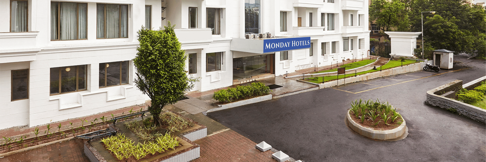 The exterior of the modern white hotel building at Monday Hotels Mahape, Navi Mumbai, with a landscaped driveway.