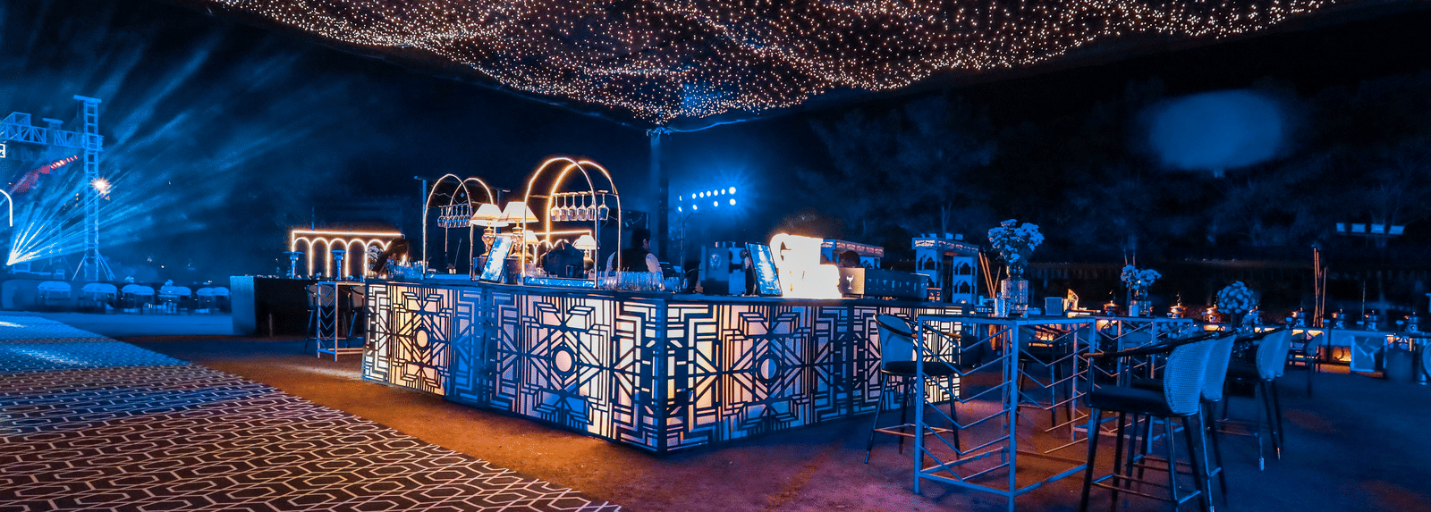 An outdoor event space at Ananta Spa & Resort, Jaipur, is illuminated by a dense canopy of warm fairy lights over a clear bar.