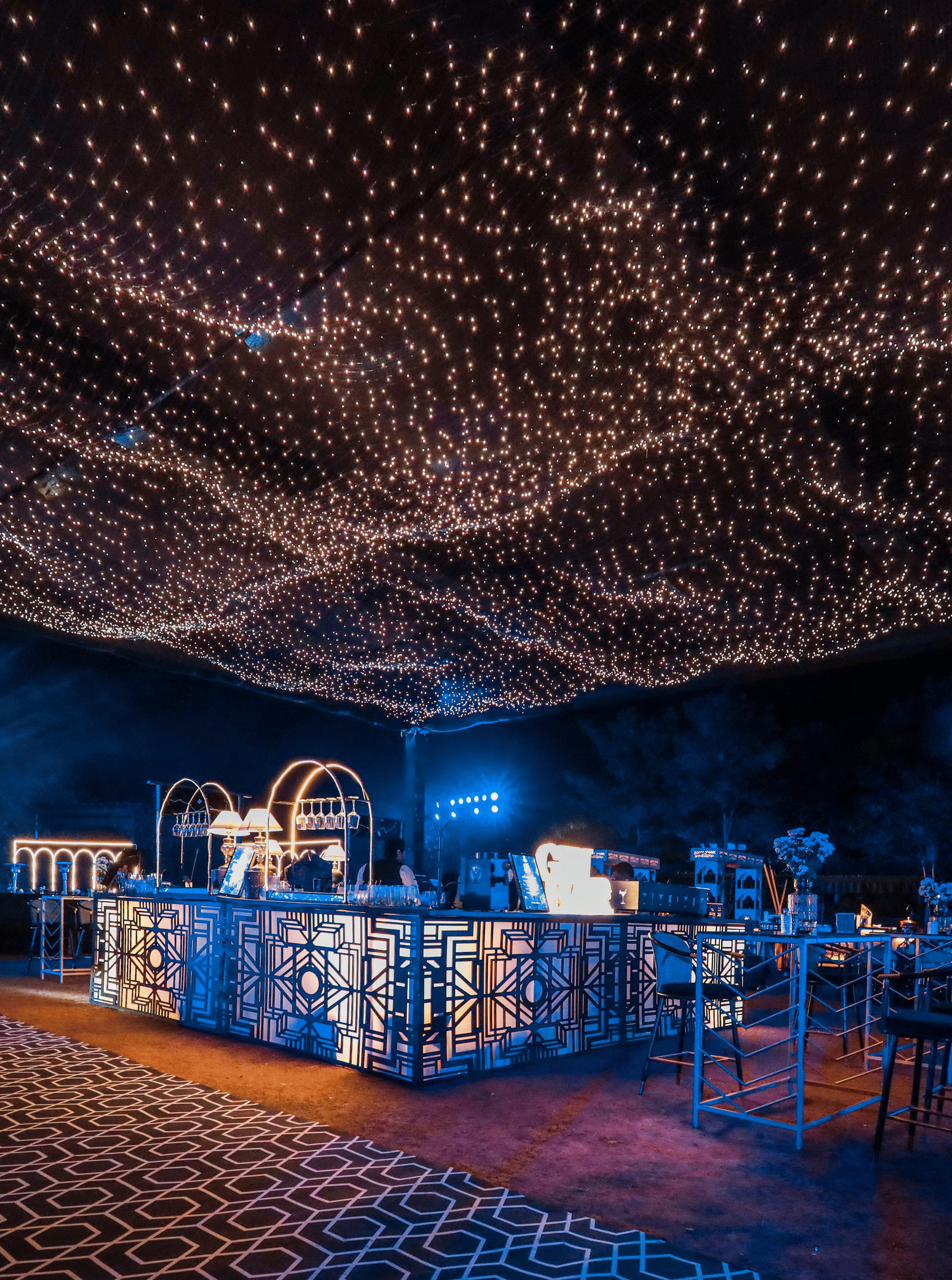 An outdoor event space at Ananta Spa & Resort, Jaipur, is illuminated by a dense canopy of warm fairy lights over a clear bar.