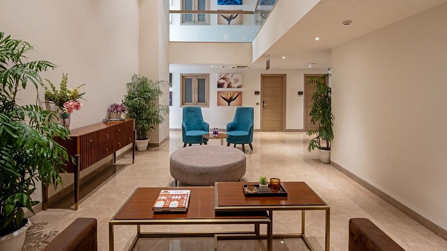 Zoomed-Out Lobby View: A wide-angle view of Hotel Saket 27’s lobby showcasing a brown table and a matching brown sofa, with two blue chairs in the background. The space is elegantly framed by plants in the corners, adding a touch of greenery to the modern 