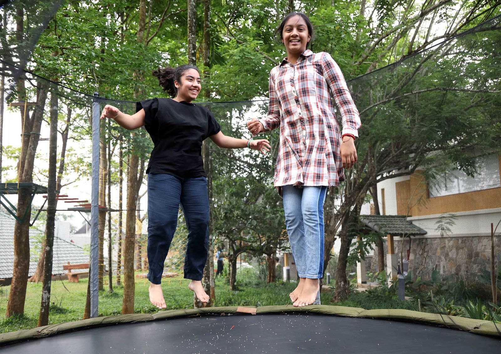 Two cheerful girls bouncing on a trampoline in a green, wooded outdoor area with cottages in the background—highlighting the playful and welcoming vibe of a child-friendly hotel in Wayanad.
