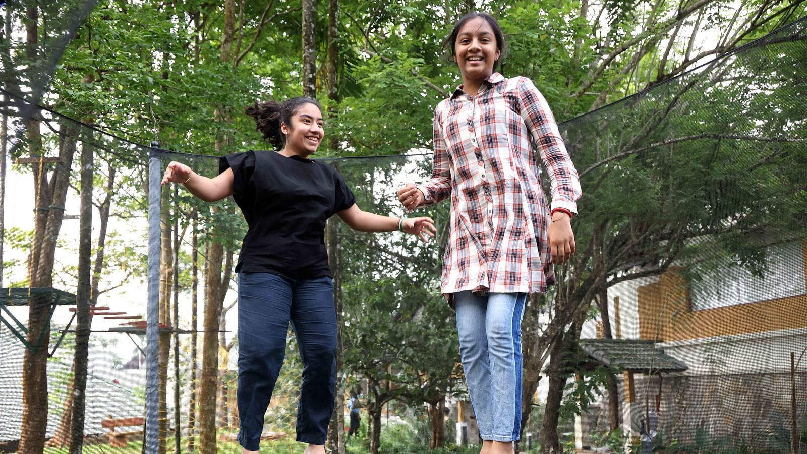 Two cheerful girls bouncing on a trampoline in a green, wooded outdoor area with cottages in the background—highlighting the playful and welcoming vibe of our child-friendly hotel in Wayanad.