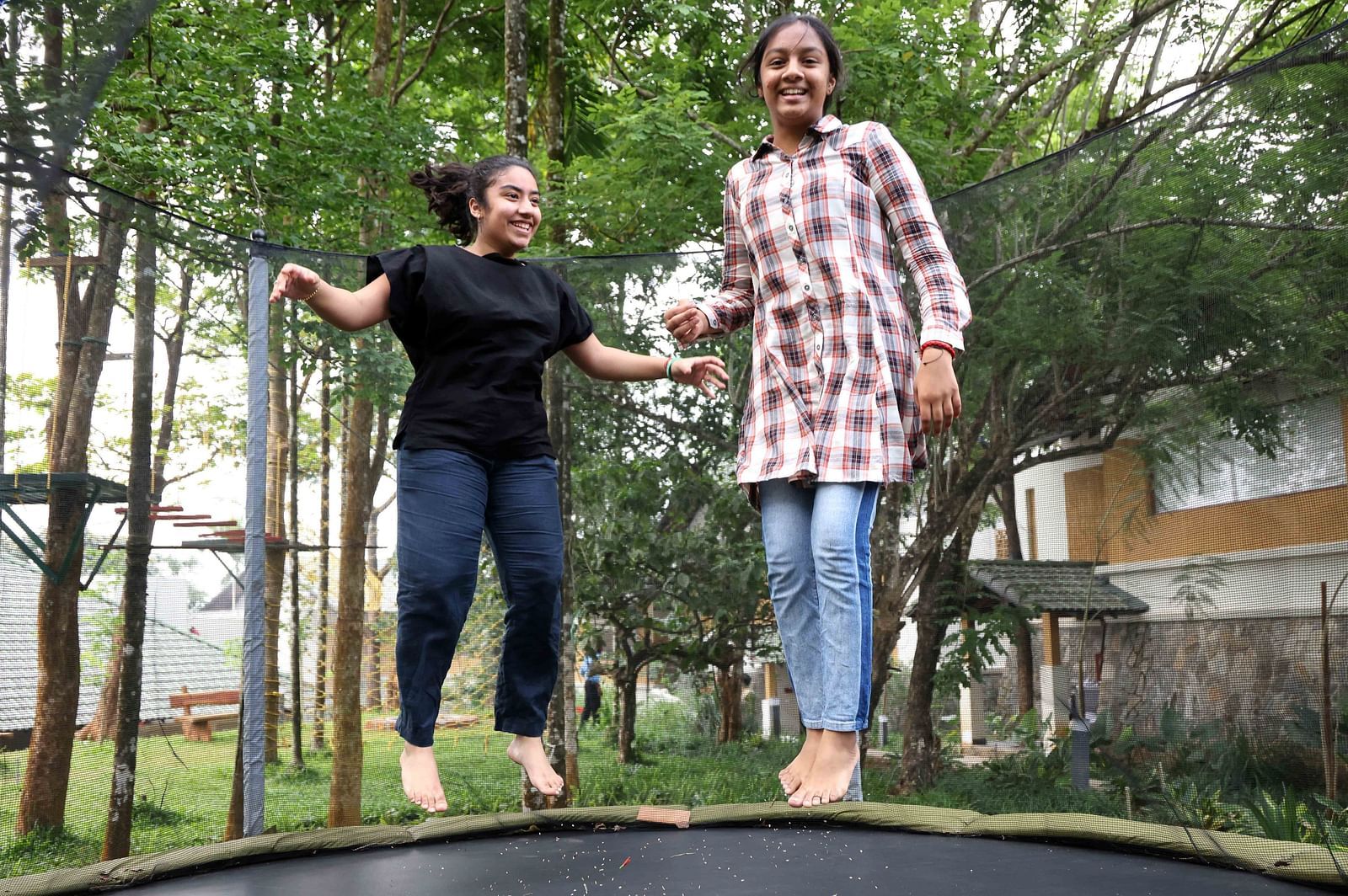 Two cheerful girls bouncing on a trampoline in a green, wooded outdoor area with cottages in the background—highlighting the playful and welcoming vibe of our child-friendly hotel in Wayanad.