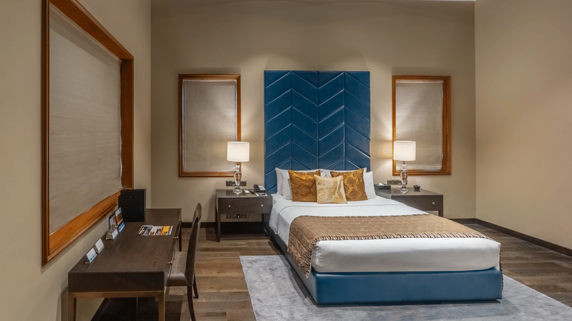 A bed flanked by bedside lamps, side tables. A large headboard and a study table with a chair are also in view at the Presidential Suite inside Kenilworth Hotel, Kolkata - A hotel near City Centre 1, Kolkata.