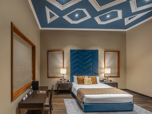 A bed flanked by bedside lamps, side tables. A large headboard and a study table with a chair are also in view at the Presidential Suite inside Kenilworth Hotel, Kolkata - A hotel near City Centre 1, Kolkata.