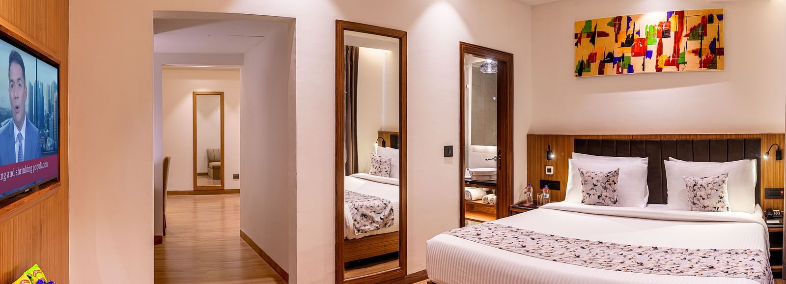 Hotel room with a king size bed, flat-screen TV, and a hallway leading to another room. It features a unique tray ceiling with lighting, a ceiling fan, and colourful abstract artwork - Lords Eco Inn Dehradun