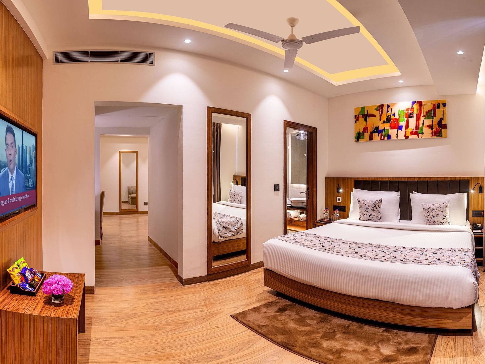 Hotel room with a king size bed, flat-screen TV, and a hallway leading to another room. It features a unique tray ceiling with lighting, a ceiling fan, and colourful abstract artwork - Lords Eco Inn Dehradun