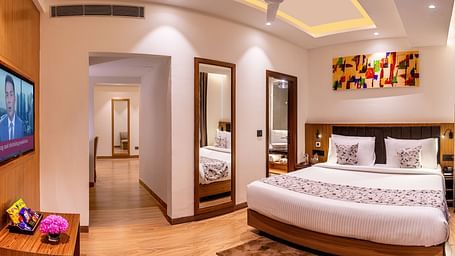 Hotel room with a king size bed, flat-screen TV, and a hallway leading to another room. It features a unique tray ceiling with lighting, a ceiling fan, and colourful abstract artwork - Lords Eco Inn Dehradun