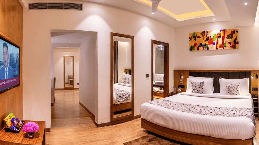 Panoramic view of a room with a king size bed, flat-screen TV, a wall art, a full-length mirror and a hallway leading to another room at Lords Eco Inn Dehradun.